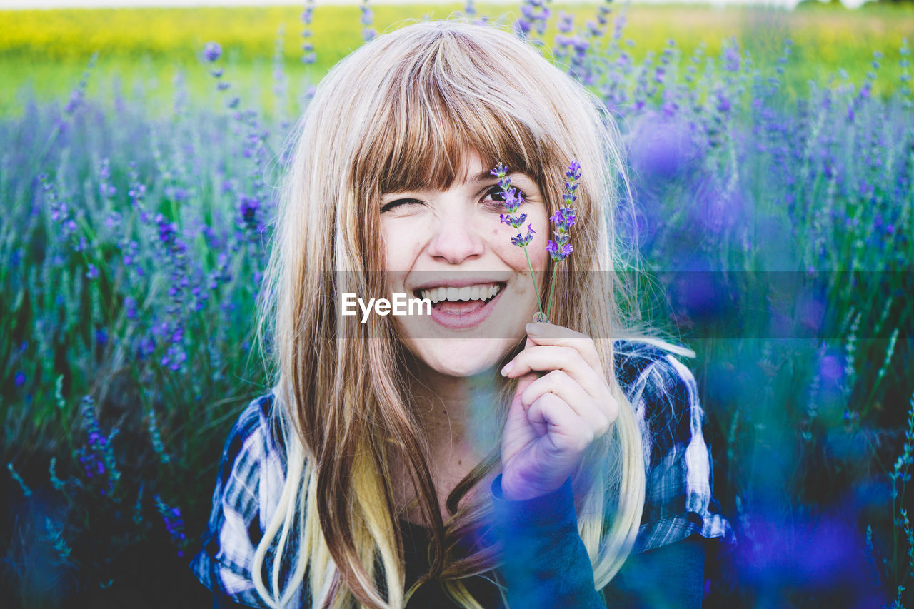 Smiling young woman holding lavender flowers over eye while sitting in field