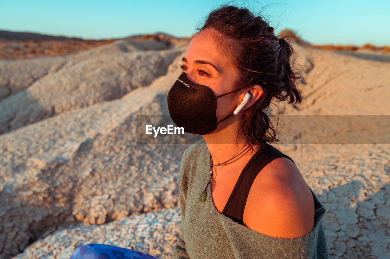 Side view of confident young sportswoman in black protective mask listening to music with wireless earbuds while recreating during training in rough desert badlands looking away