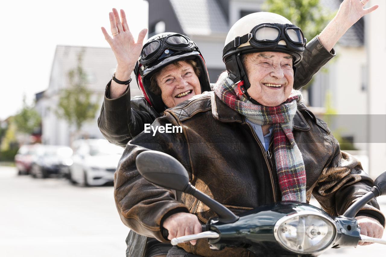 Happy senior couple having fun, riding motor scooter and waving