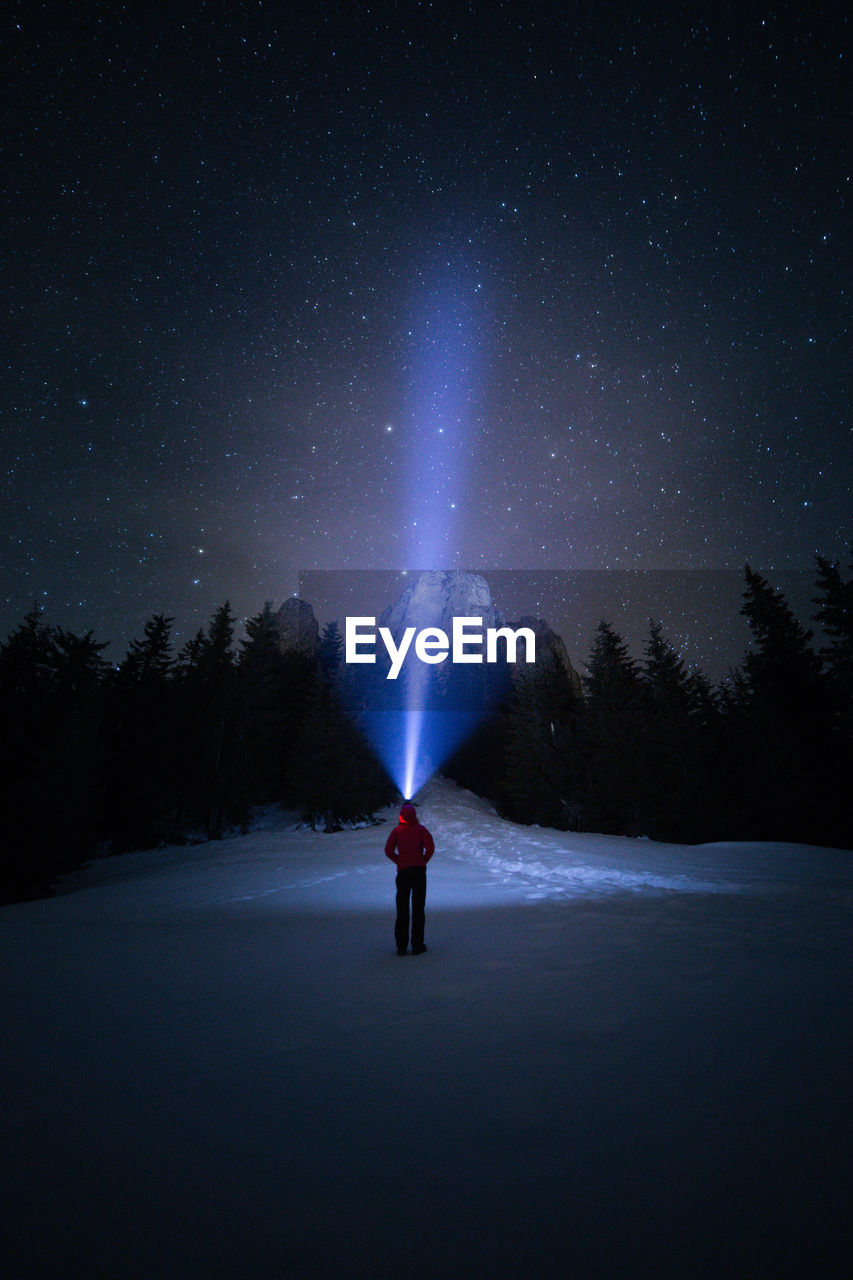 Rear view of person holding illuminated flashlight while standing on snow covered field against sky at night