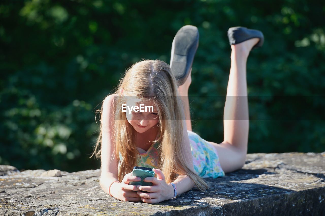 Girl using smart phone while lying on retaining wall outdoors