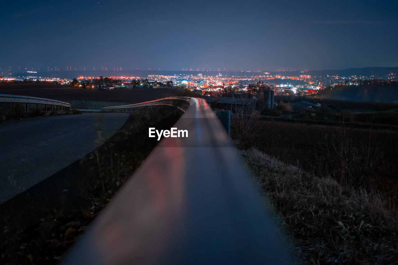 Light trails on road by illuminated city against sky at night