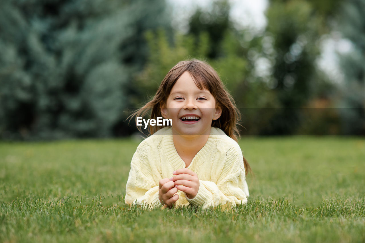 Funny smiling blonde little child girl 4-5 year old lying on green grass lawn outdoor. 