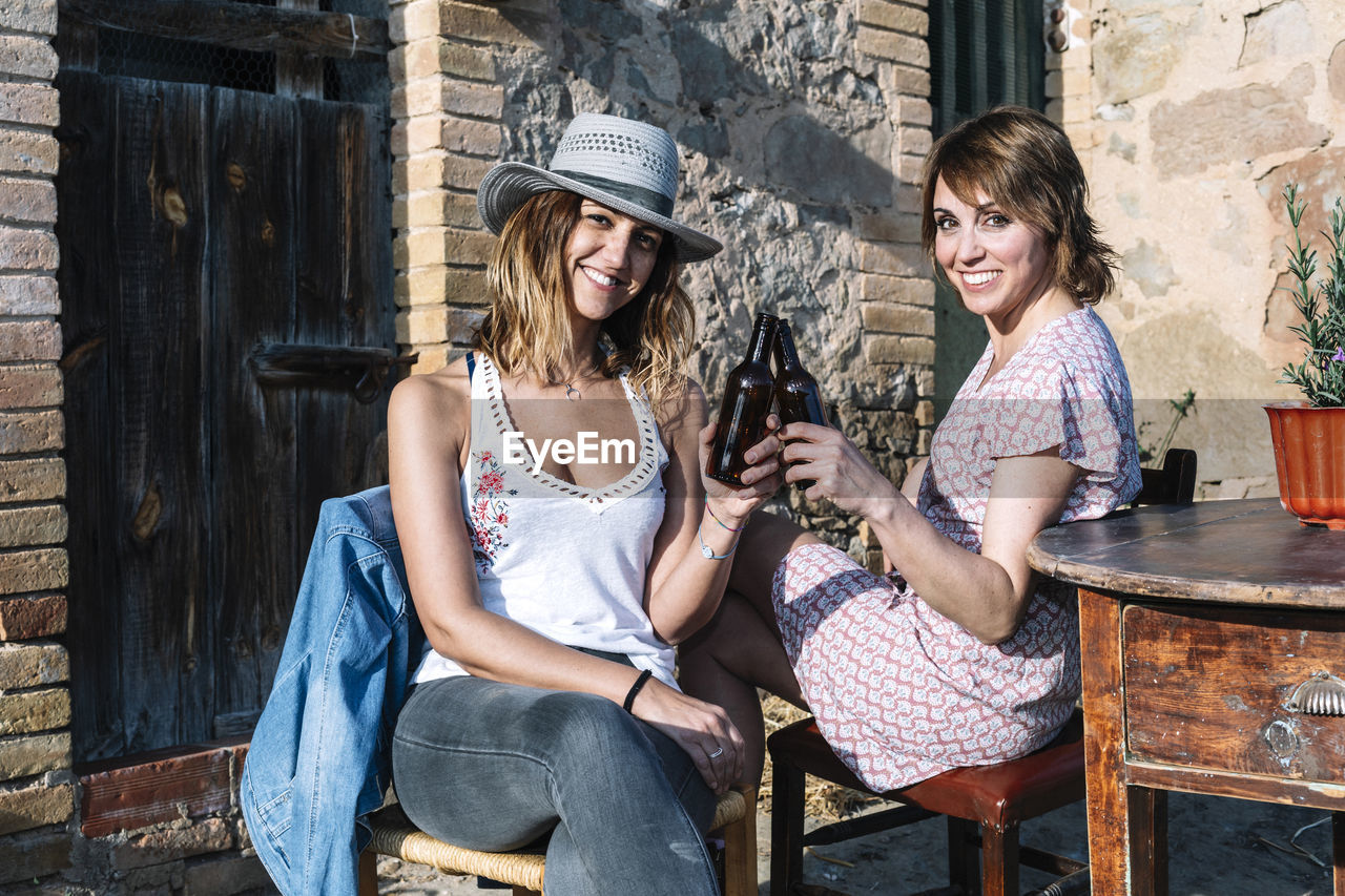 Portrait of smiling female friends doing celebratory toast against brick wall