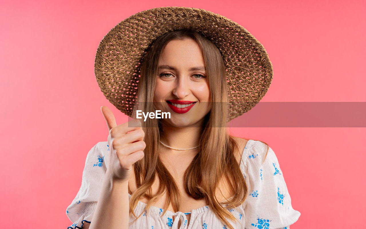 portrait, colored background, one person, smiling, hat, women, adult, clothing, happiness, studio shot, young adult, pink, looking at camera, emotion, sun hat, human face, long hair, pink background, headshot, hairstyle, front view, straw hat, cheerful, fashion, fashion accessory, indoors, fun, blond hair, human hair, positive emotion, sombrero, casual clothing, person, waist up, teeth, nose, lifestyles, smile, female, enjoyment, brown hair