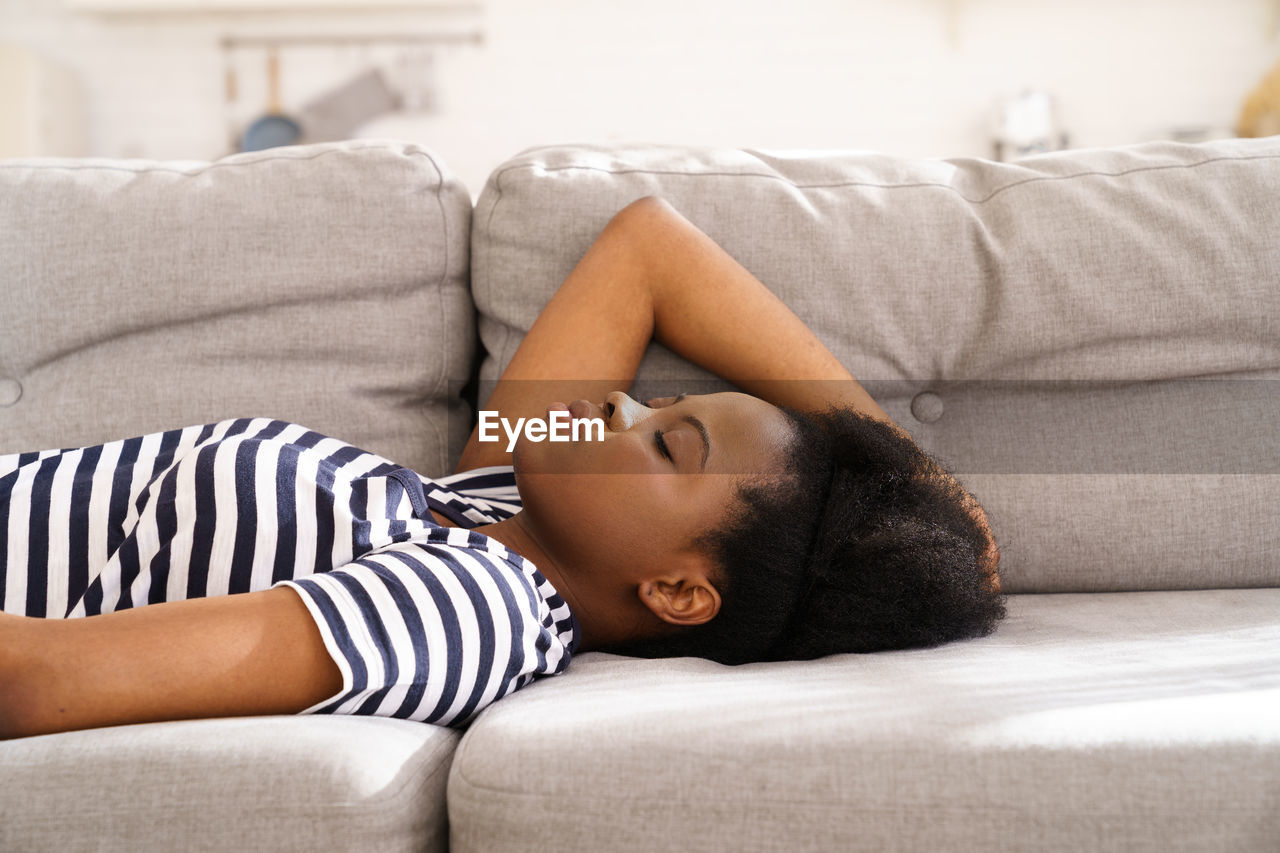 Black young woman wear stripped t-shirt resting sleeping on couch at home closed eyes taking a break
