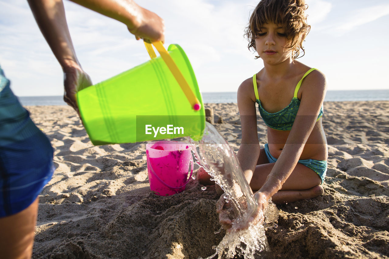 Boy pouring water on sister's hands while making sand castle at beach