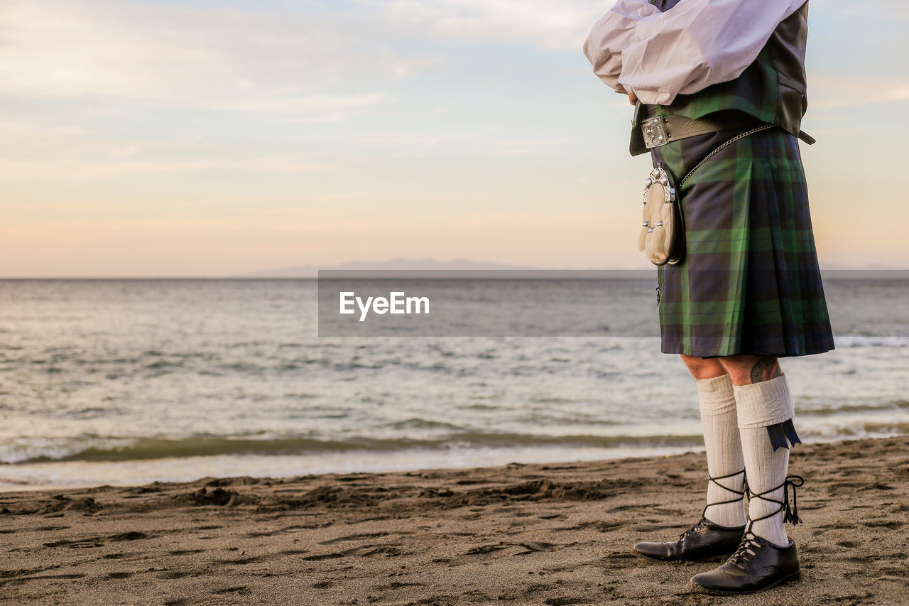 LOW SECTION OF WOMAN STANDING AT BEACH AGAINST SKY