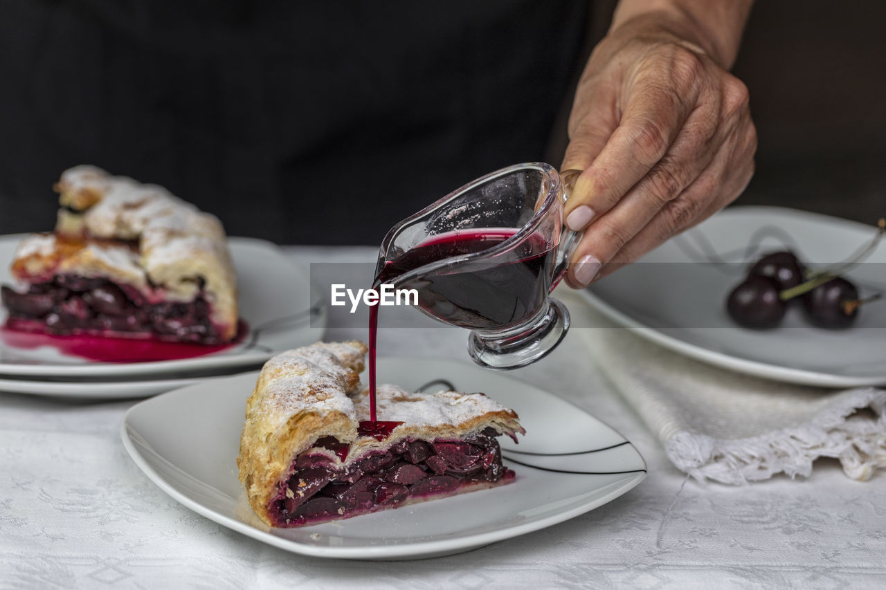 A female hand pours cherry syrup over a slice of cherry pie in the foreground. 