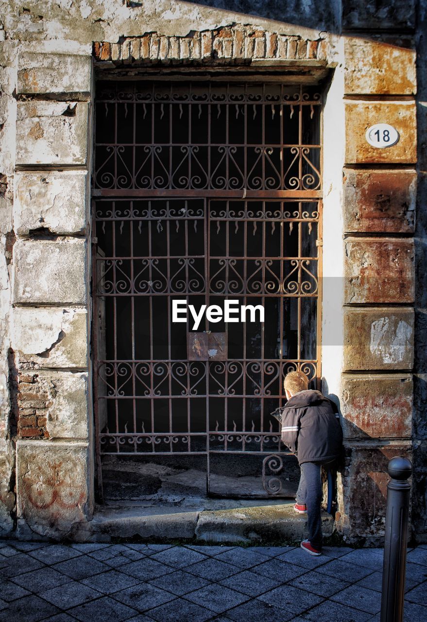 Rear view of boy wearing warm clothing peeking through gate in city