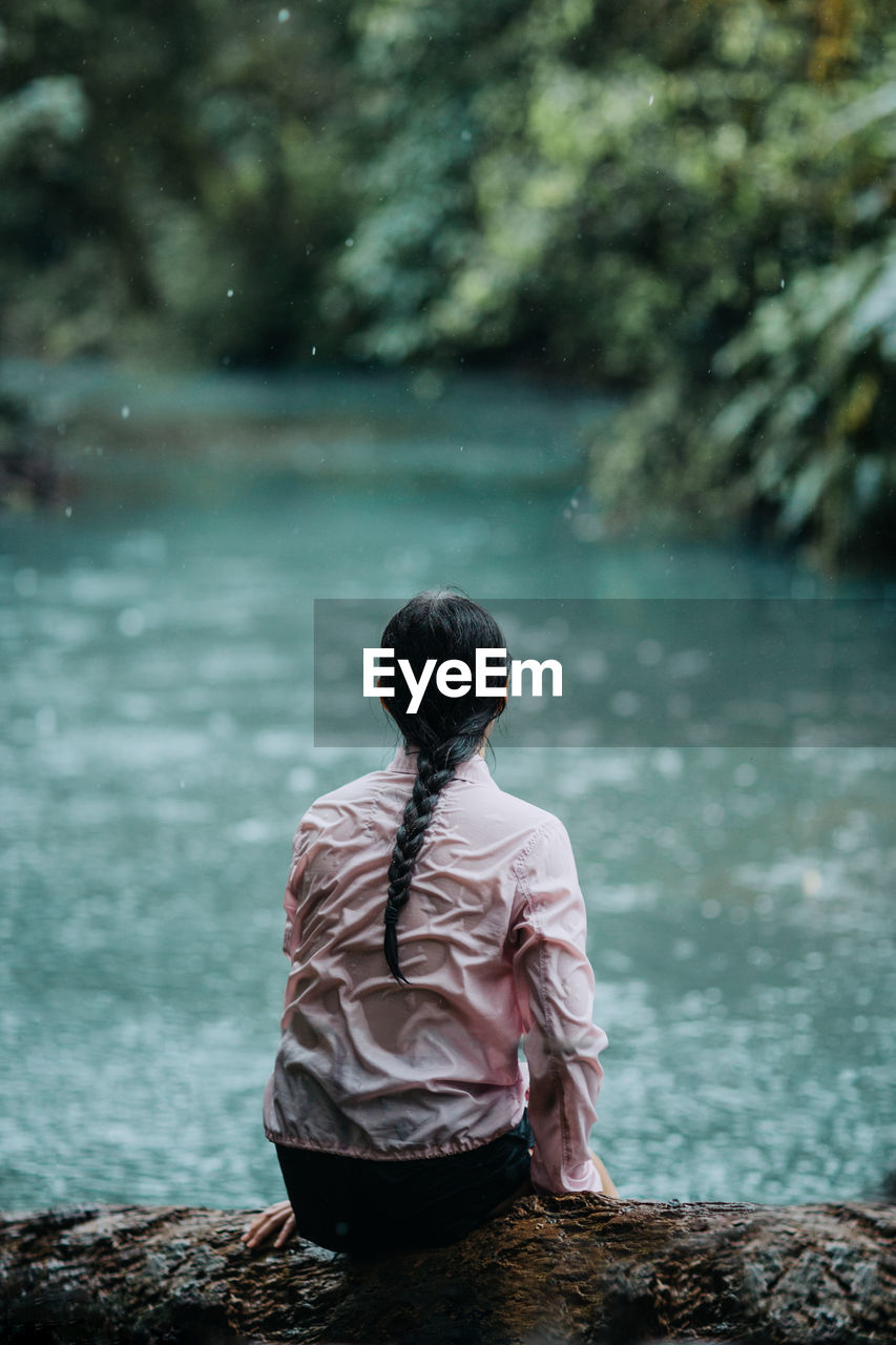 Back view of unrecognizable female traveler sitting on shore of calm celeste river with turquoise water flowing through green rainforest while relaxing and enjoying solitude during summer journey in costa rica