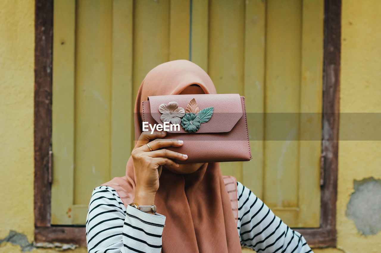 Woman holding purse over face against wall
