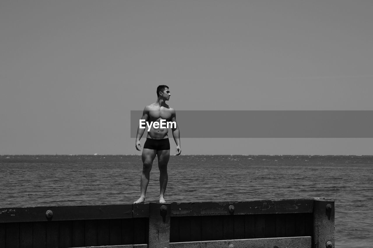 FULL LENGTH OF SHIRTLESS MAN STANDING ON PIER AGAINST SEA