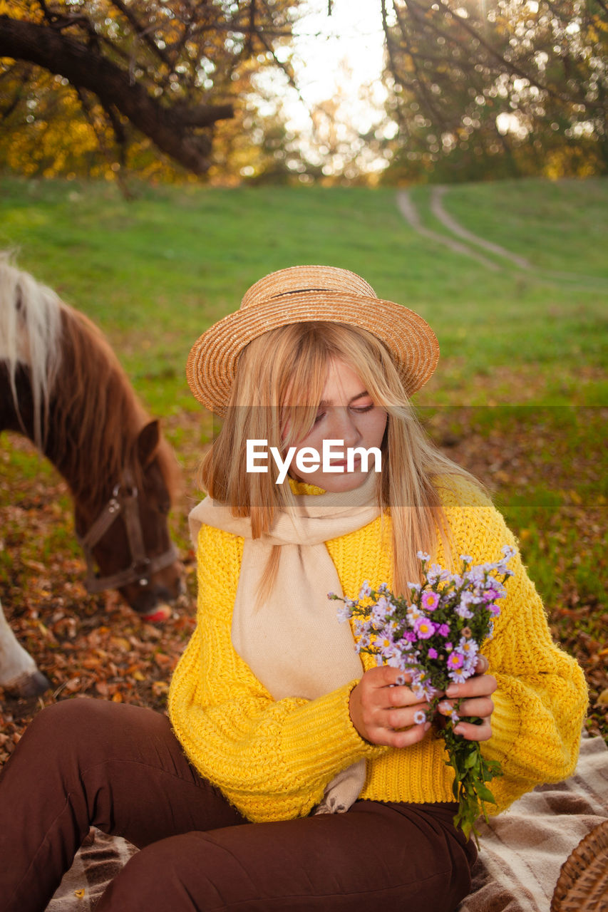 yellow, women, autumn, plant, adult, nature, one person, clothing, blond hair, flower, smiling, flowering plant, happiness, emotion, long hair, beauty in nature, sitting, leisure activity, hairstyle, lifestyles, horse, young adult, female, fashion accessory, hat, tree, outdoors, rural scene, front view, landscape, three quarter length, portrait, positive emotion, field, person, relaxation, holding, spring, day, land, cheerful, casual clothing, grass, freshness, child, enjoyment, leaf, looking at camera, dress, springtime, human face, animal, animal themes