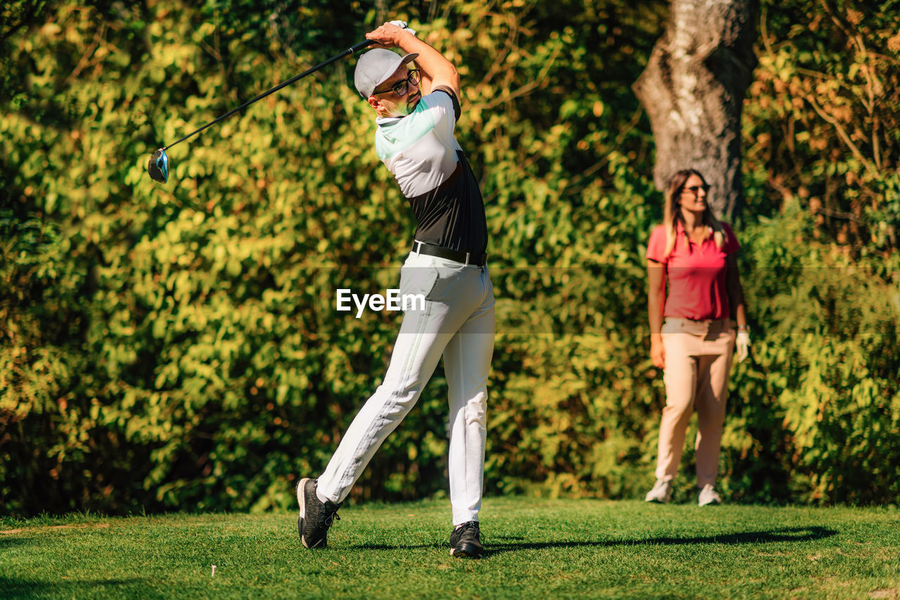 Golfing couple. man teeing off with the driver, lady in the back, following the ball in flight