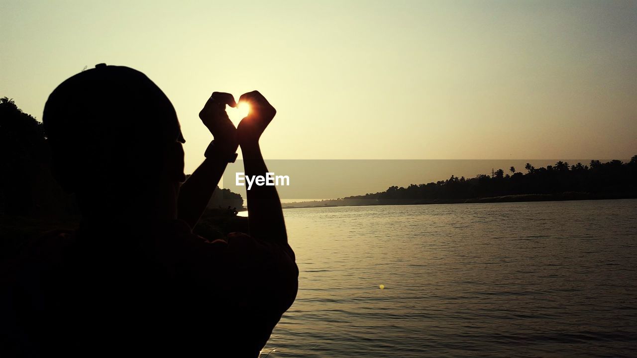 Silhouette man making heart shape at lakeshore during sunset