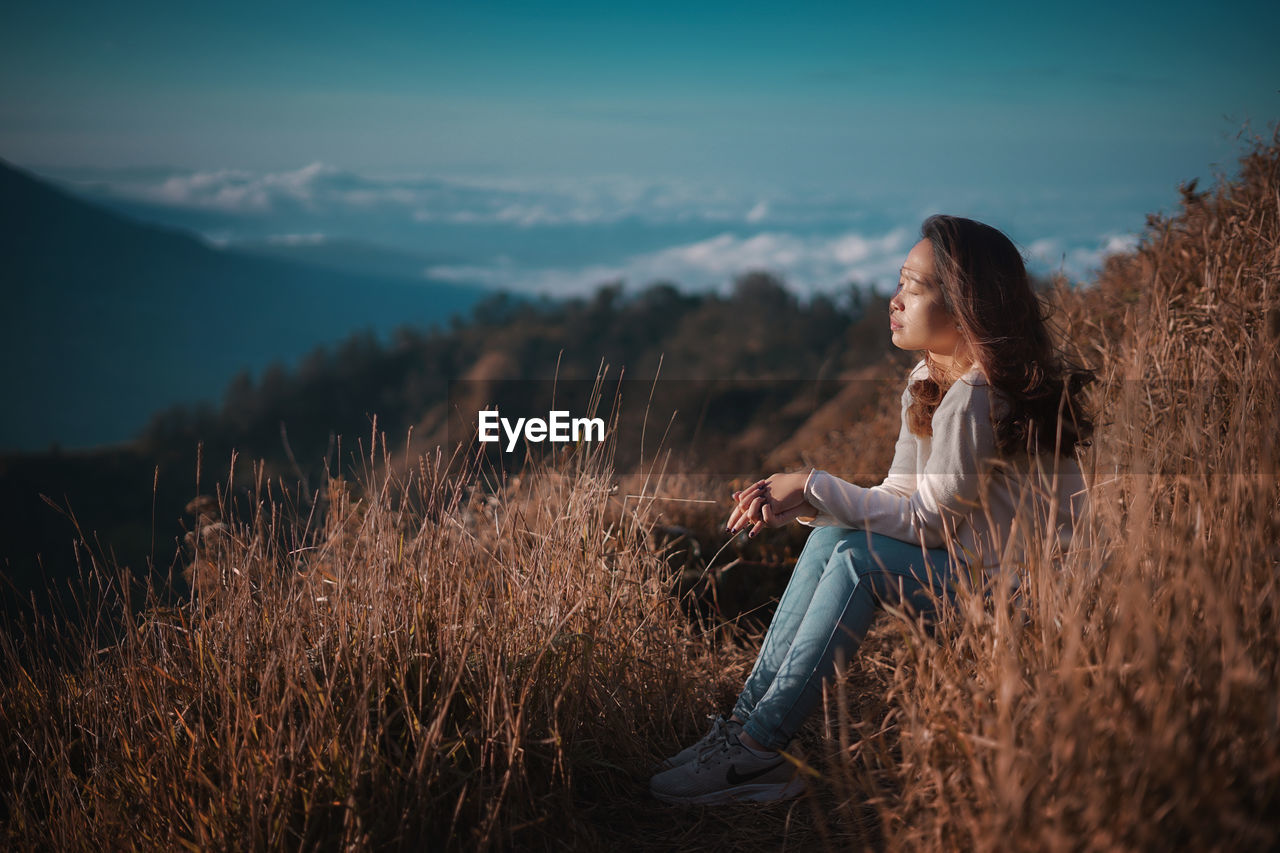 Side view of woman sitting on high field against sky