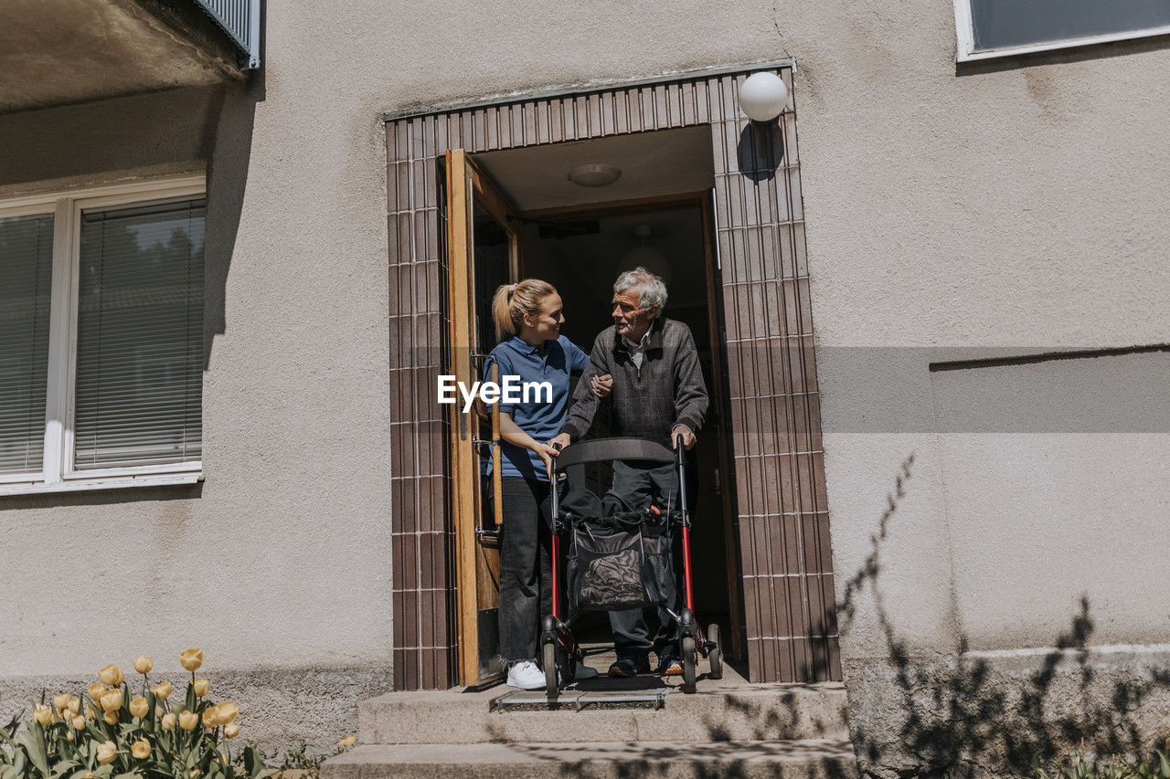 Female caregiver assisting senior man moving out with walker