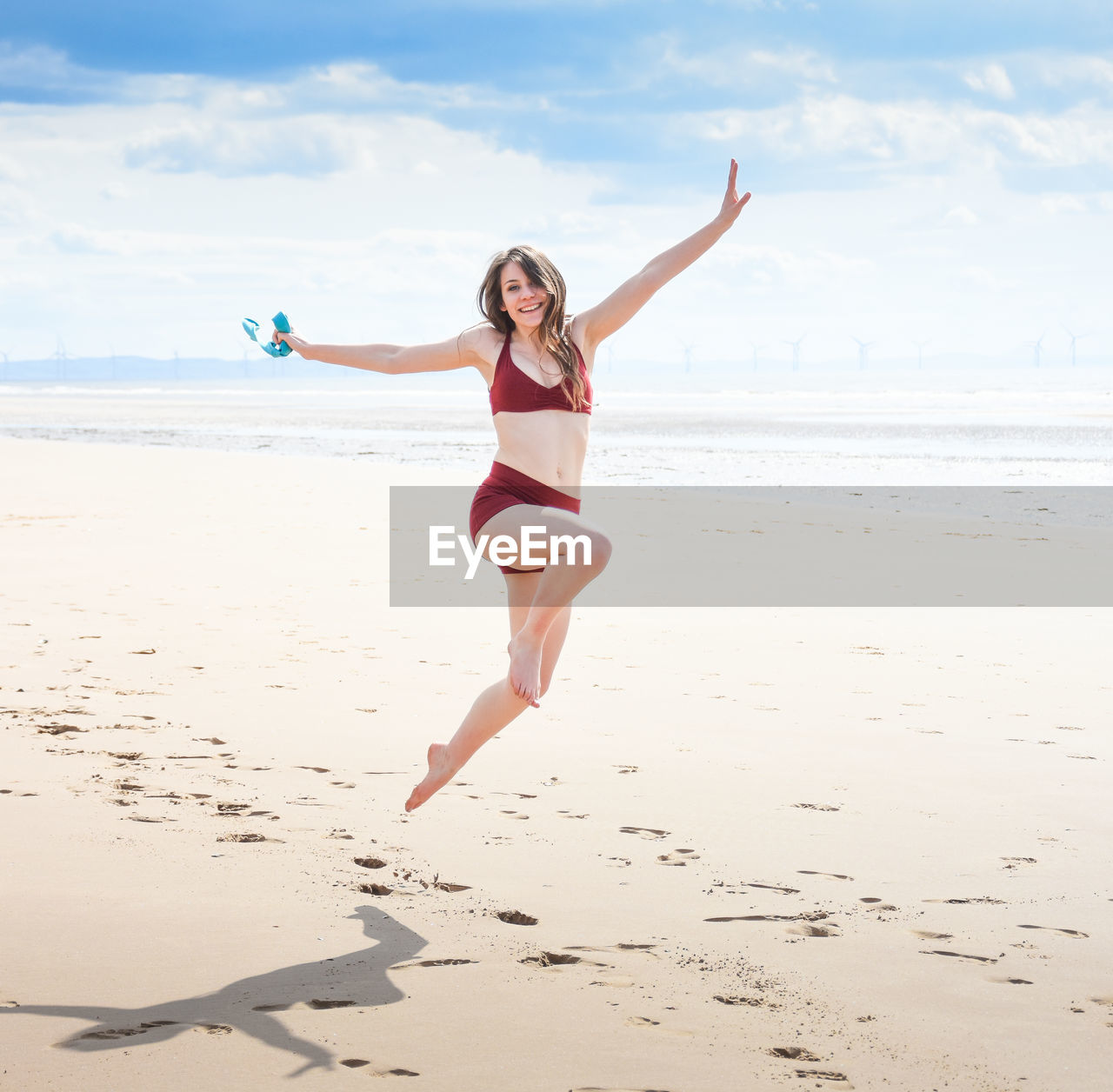 FULL LENGTH OF YOUNG WOMAN JUMPING ON BEACH AGAINST SEA