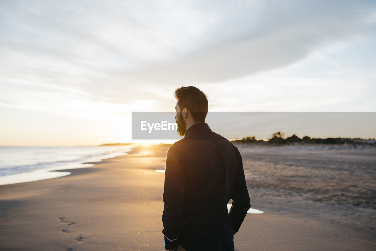 Rear view of mid adult man standing at beach against cloudy sky during sunset