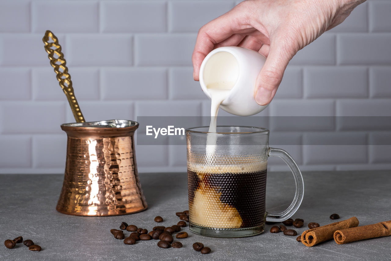 A woman's hand pours cream into a glass cup with coffee next to a traditional turkish coffee pot. 