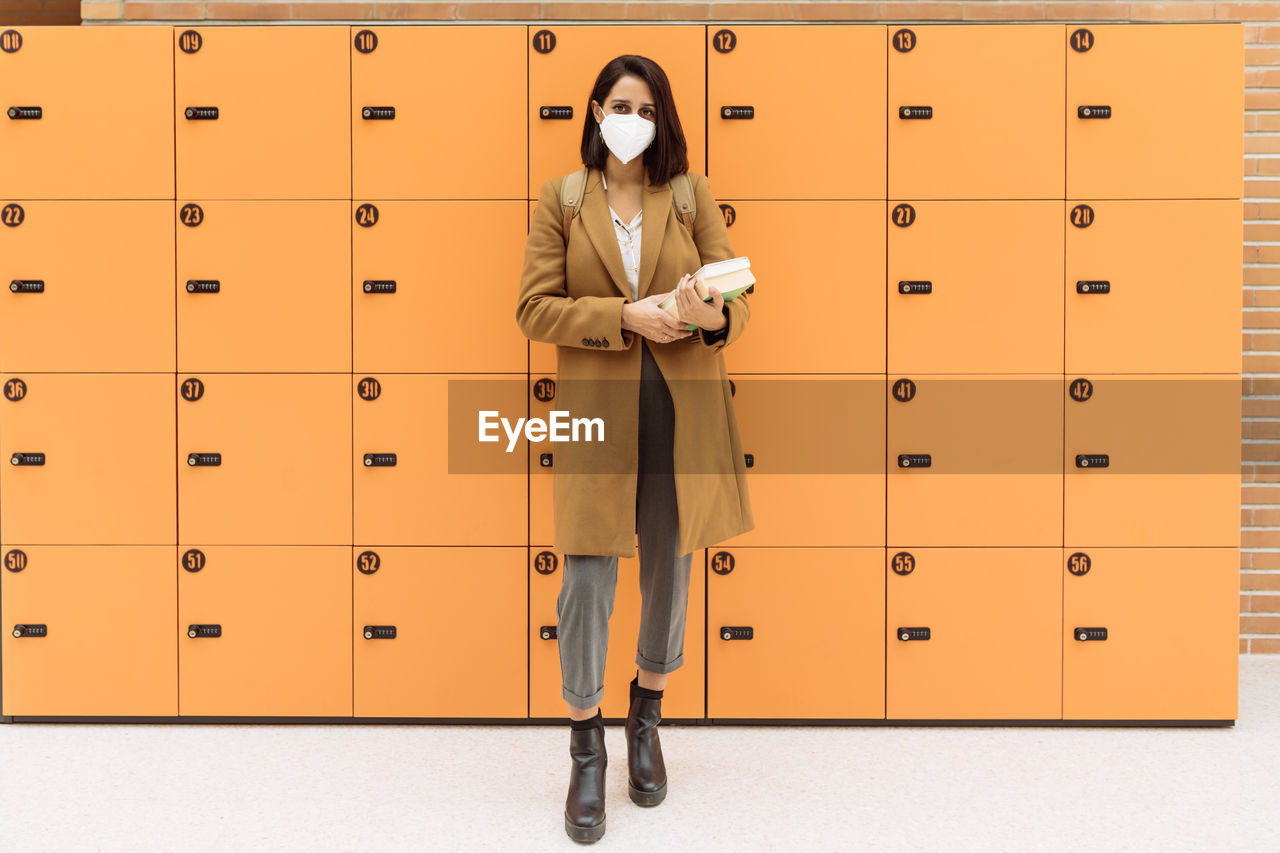 Female student wearing protective mask standing with pile of books near lockers in university corridor and looking at camera during coronavirus epidemic