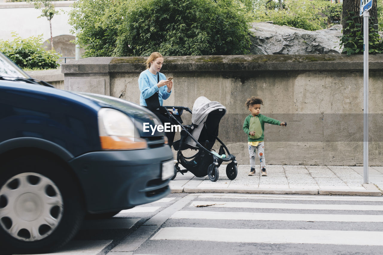 Mother using mobile phone while standing with son and baby stroller on sidewalk at city