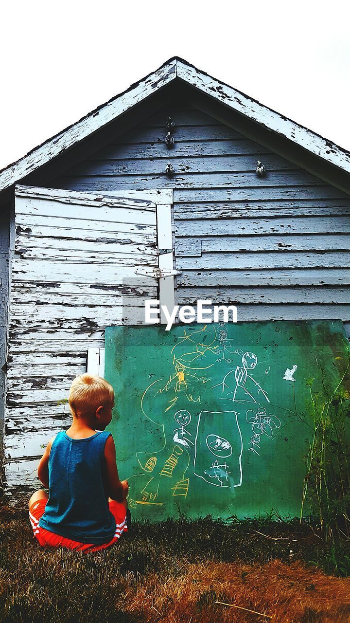 Rear view of boy chalk drawing on board while sitting at field in yard