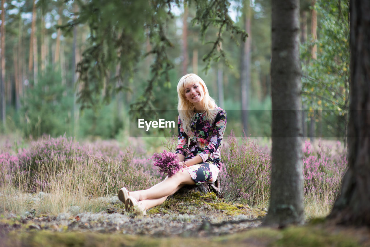 Portrait of woman holding flowers while sitting by tree trunk in forest