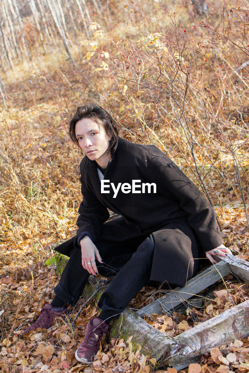 A young woman wearing a black coat sitting on a log in a serene autumn forest. 