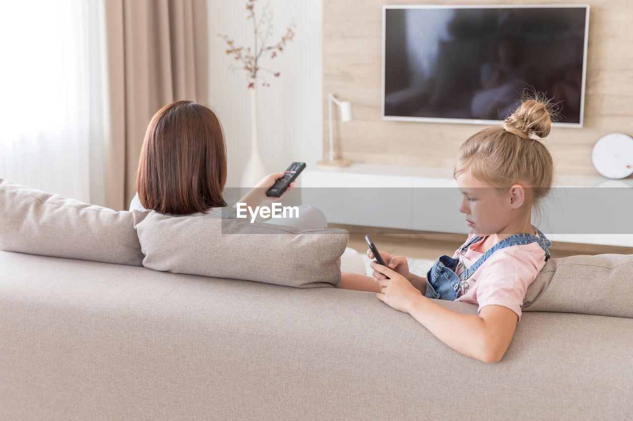 Girl using smart phone sitting by mother on sofa at home
