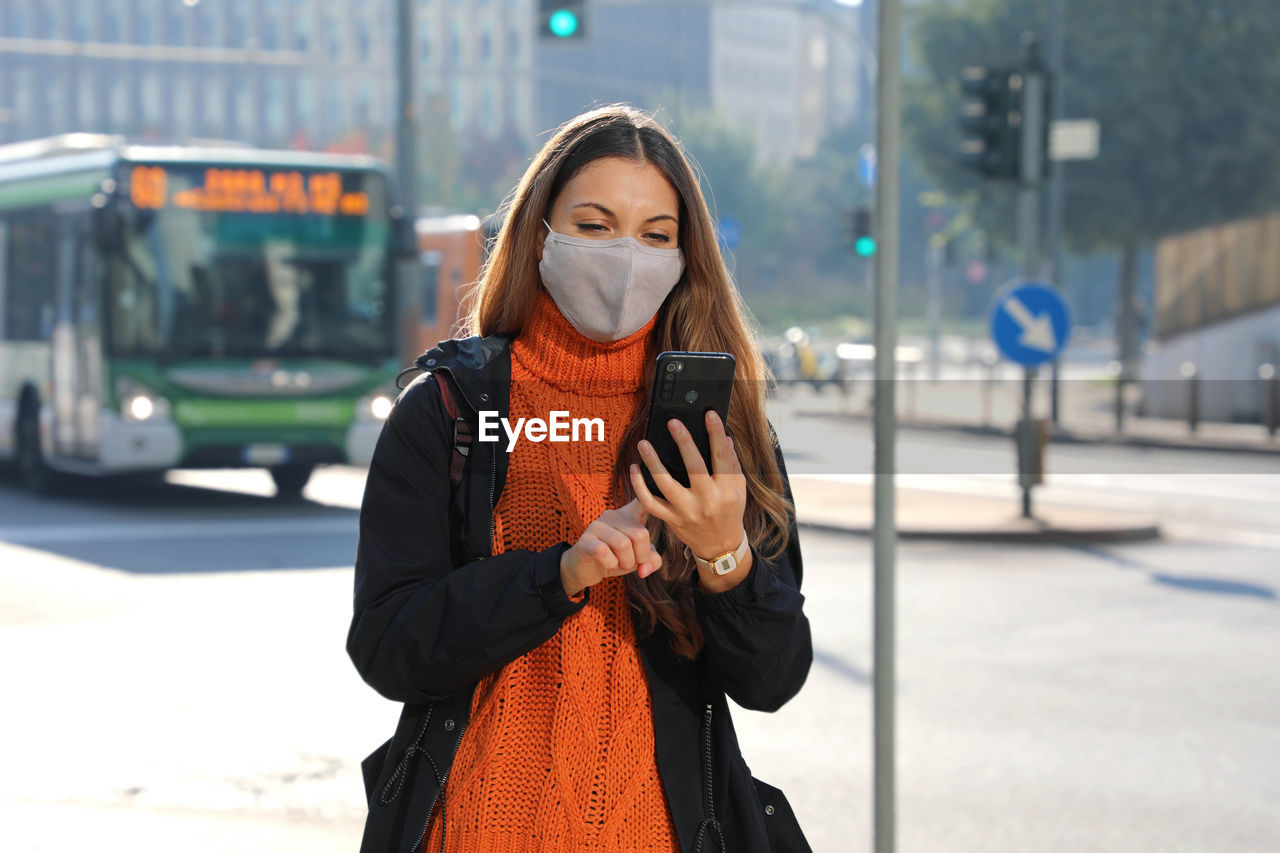 Smiling young woman wearing mask using smart phone while standing outdoors