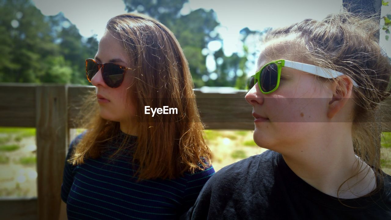 Female friends wearing sunglasses while standing by wooden fence