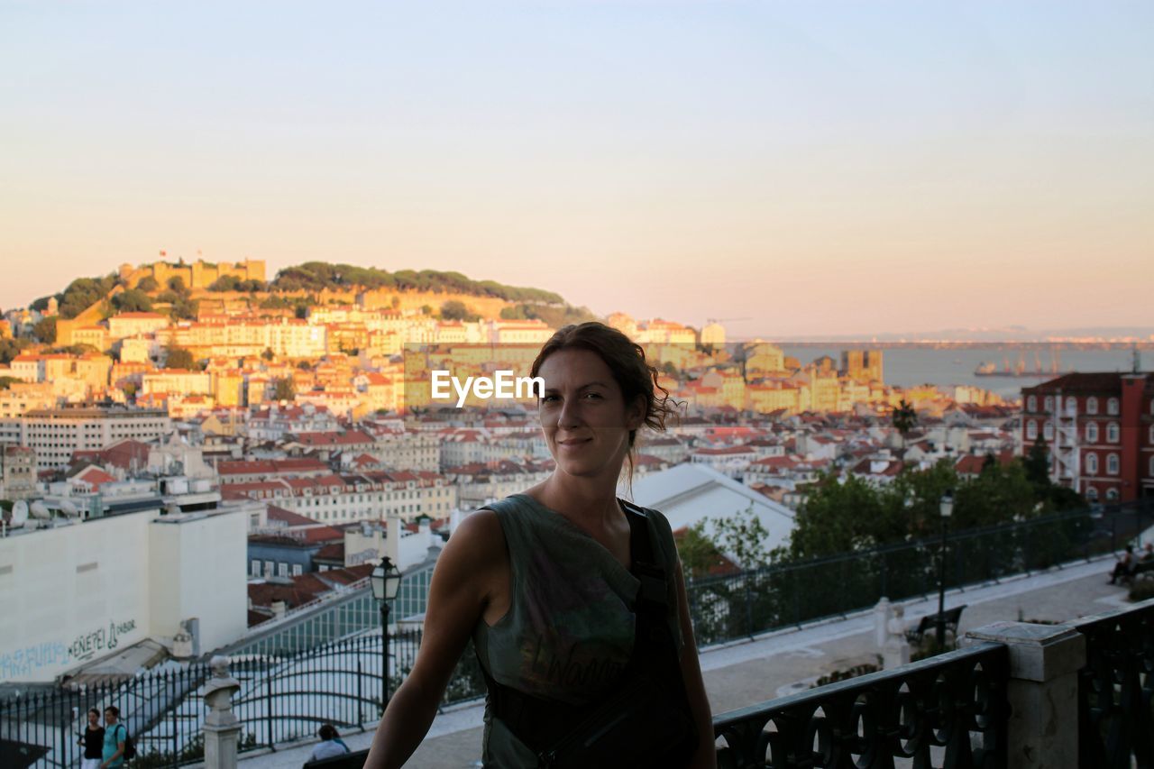 Portrait of woman standing against cityscape in balcony during sunset