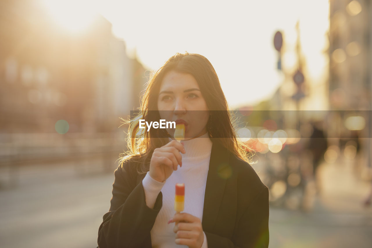 Young woman eating flavored ice while standing on road in city during sunset