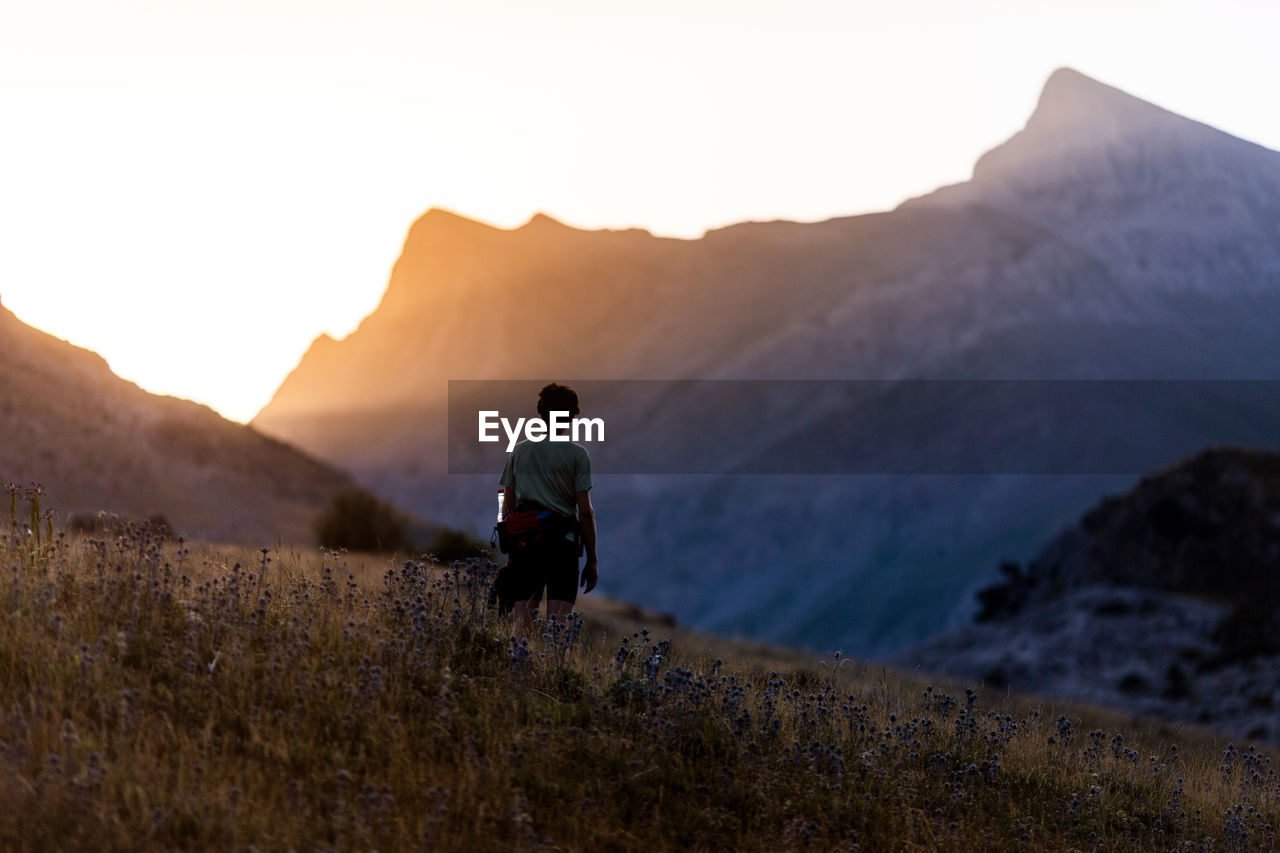 Back view of unrecognizable traveler standing in grassy valley and admiring mountain ridge at sunrise in pyrenees