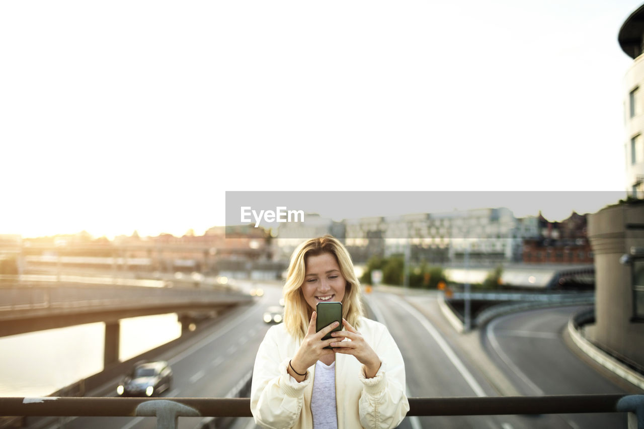 Happy young woman using mobile phone while standing on bridge against sky