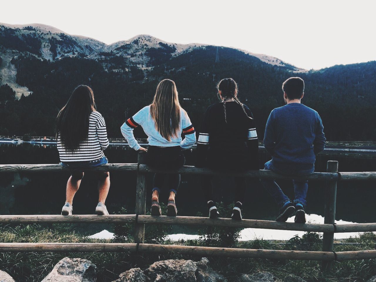 Rear view full length of friends sitting on railing by lake against mountain