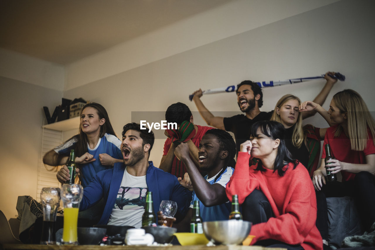 Male and female friends watching soccer match while sitting against wall at home