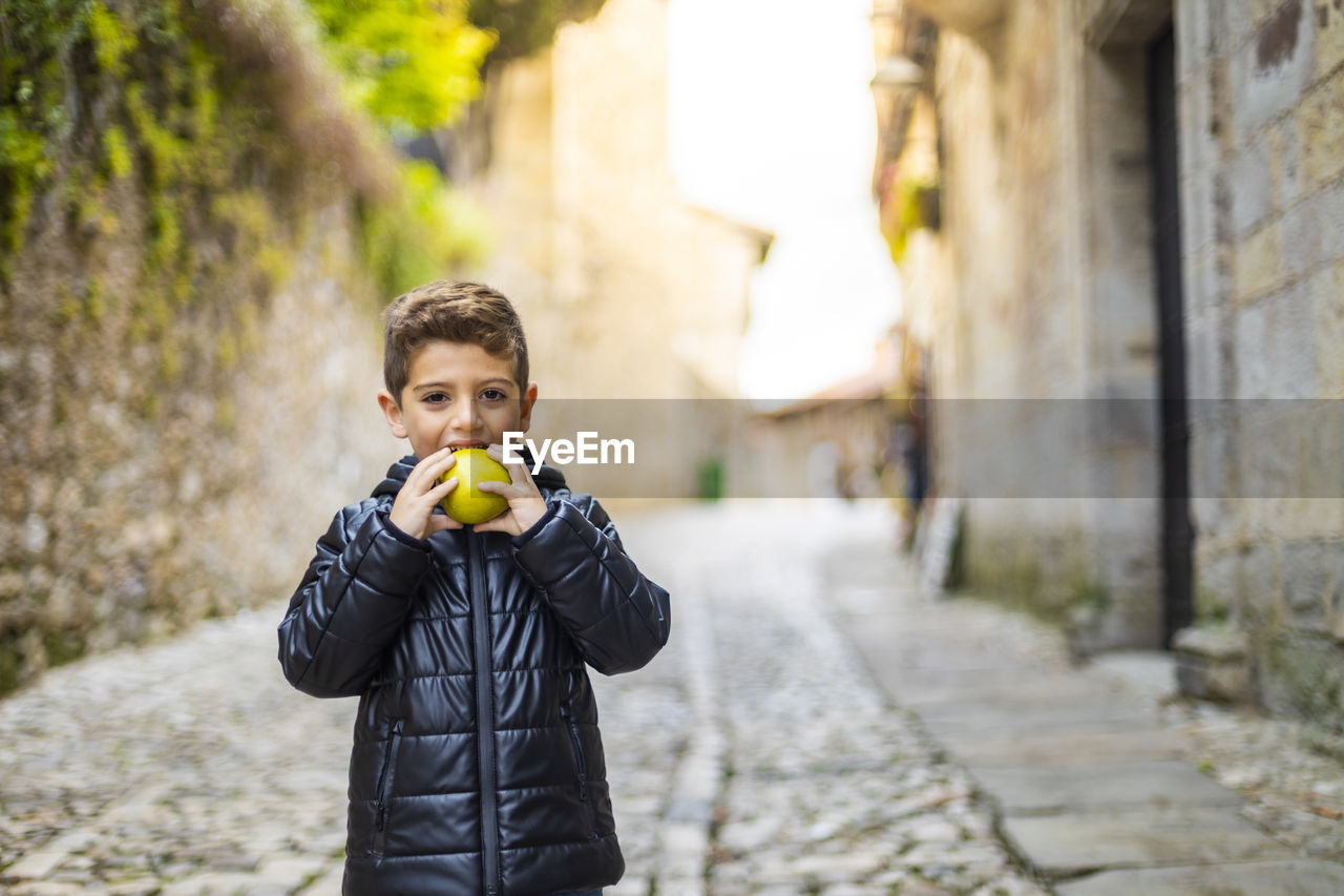 Portrait of smiling boy eating apple while standing on street