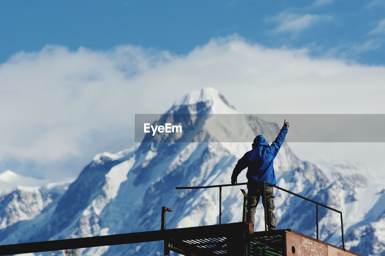 Man with arms raised standing on observation point by snowcapped mountains