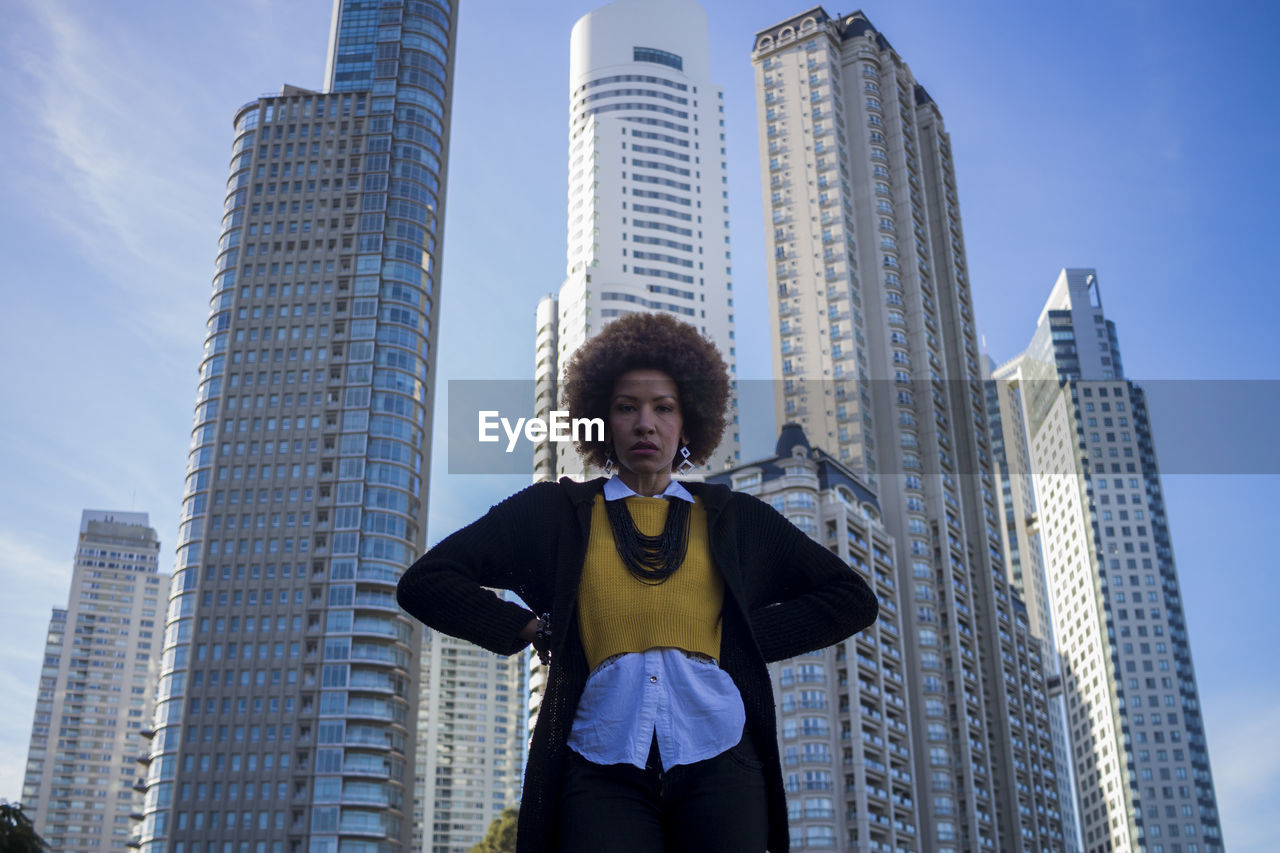 Low angle portrait of woman standing against modern buildings in city