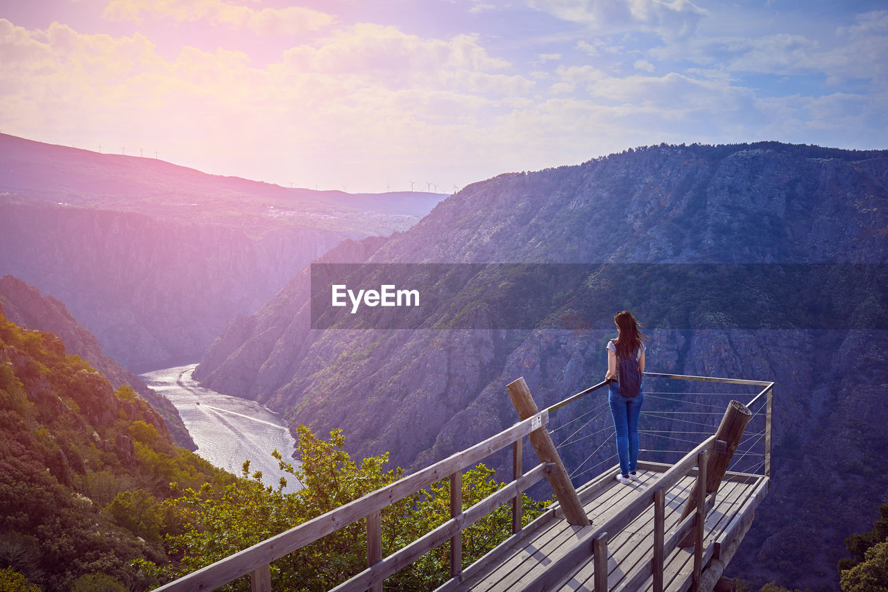 Rear view of woman standing by railing at observation point against mountain range and sky