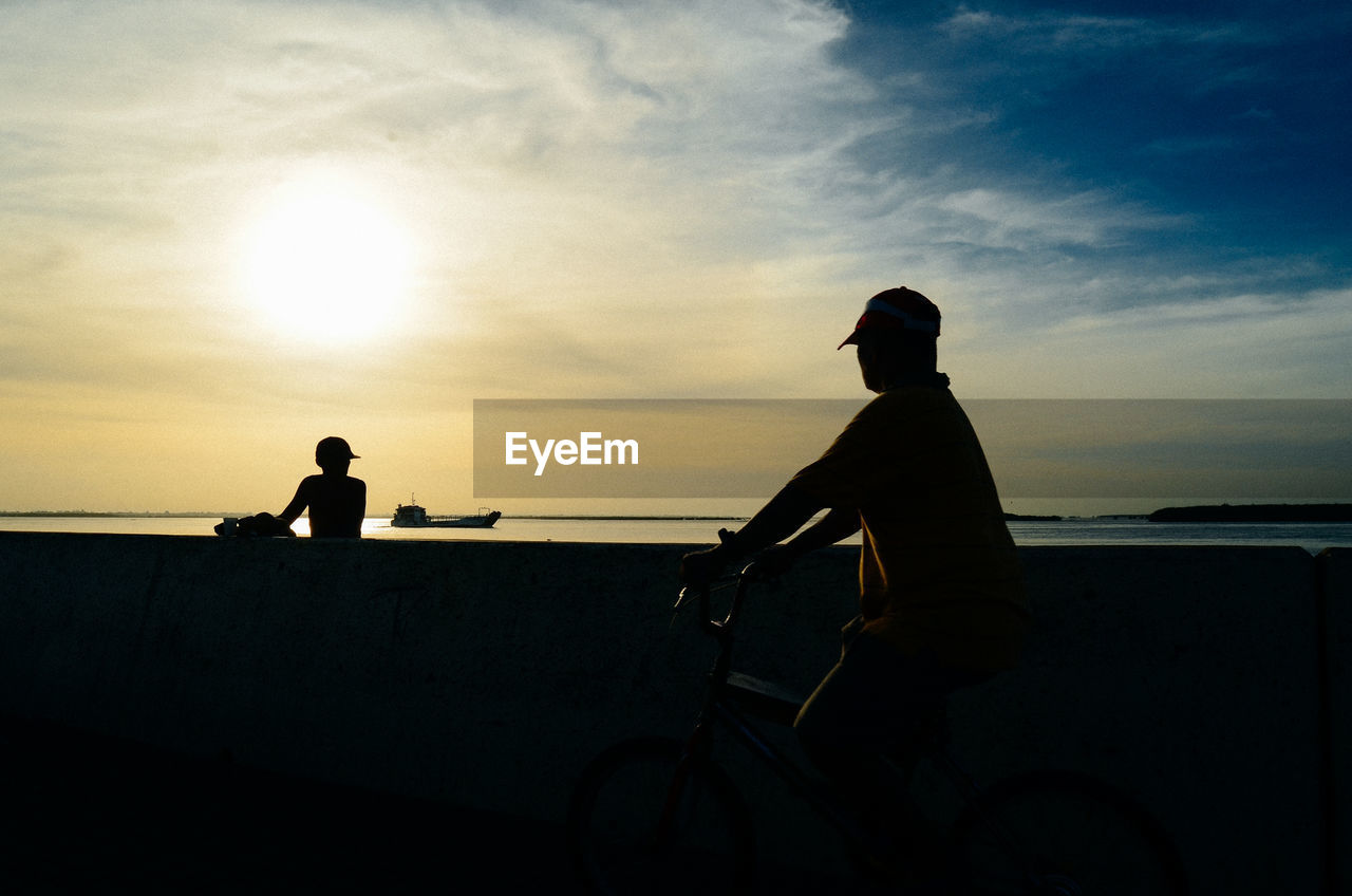 SILHOUETTE MEN SITTING ON BEACH AGAINST SKY AT SUNSET