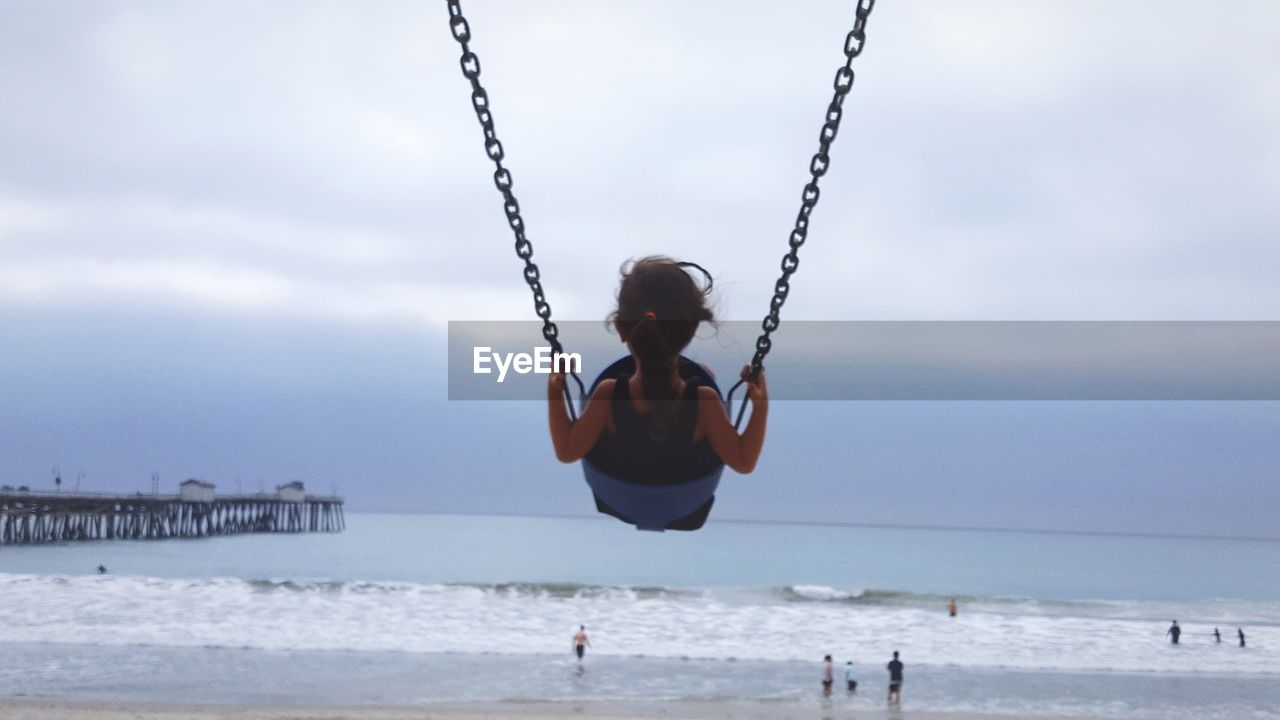 Rear view of girl playing on swing at beach against sky