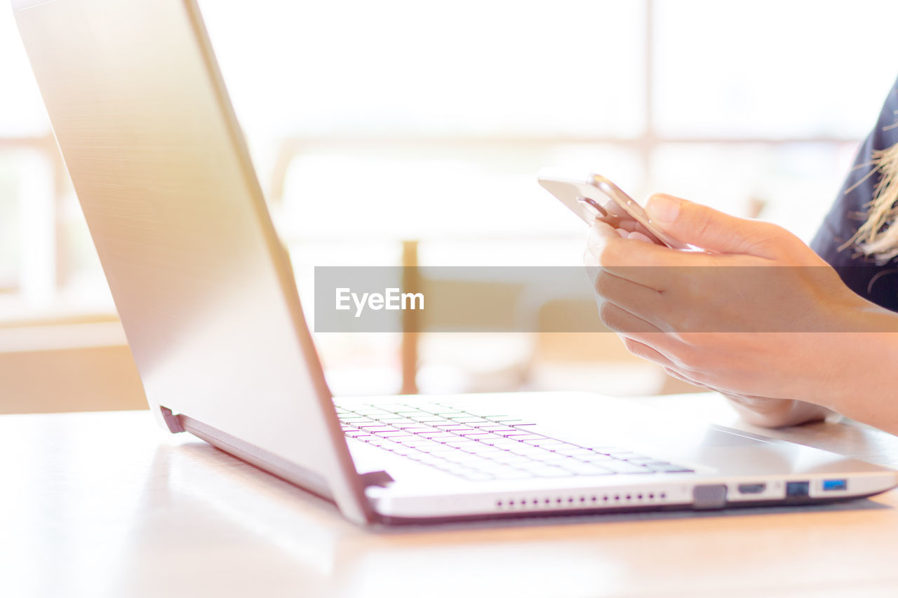 Cropped hands of woman using mobile phone by laptop at home