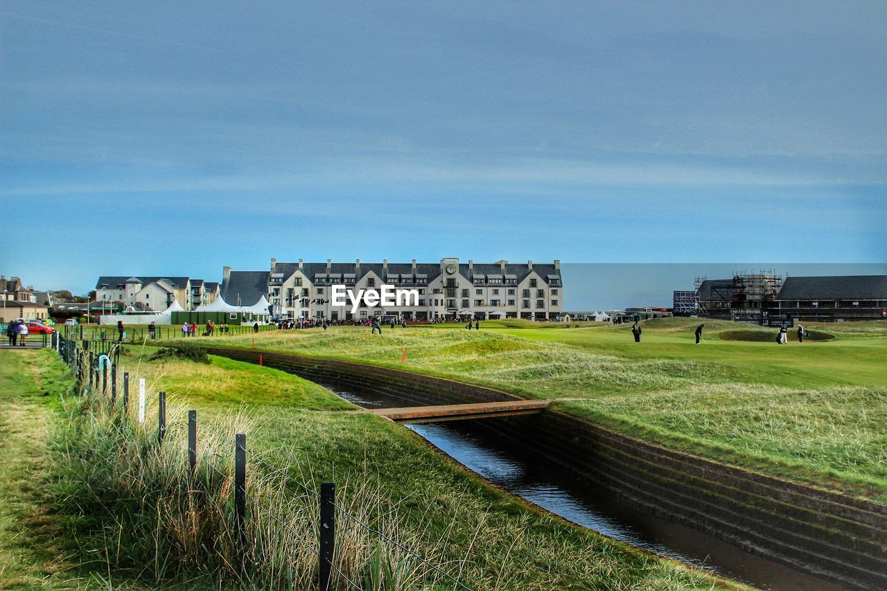 Distant view of people on grassy field by buildings against sky