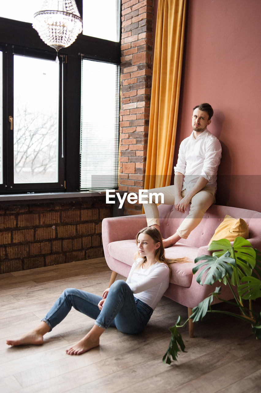 Man sits on sofa's back and look at camera, woman sits on floor near.