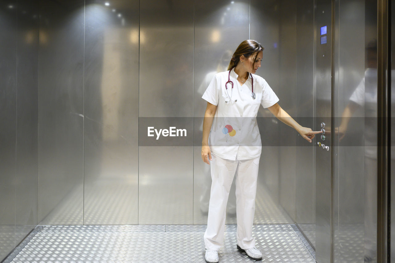 Full body woman in white medical uniform pushing button inside elevator while working in hospital