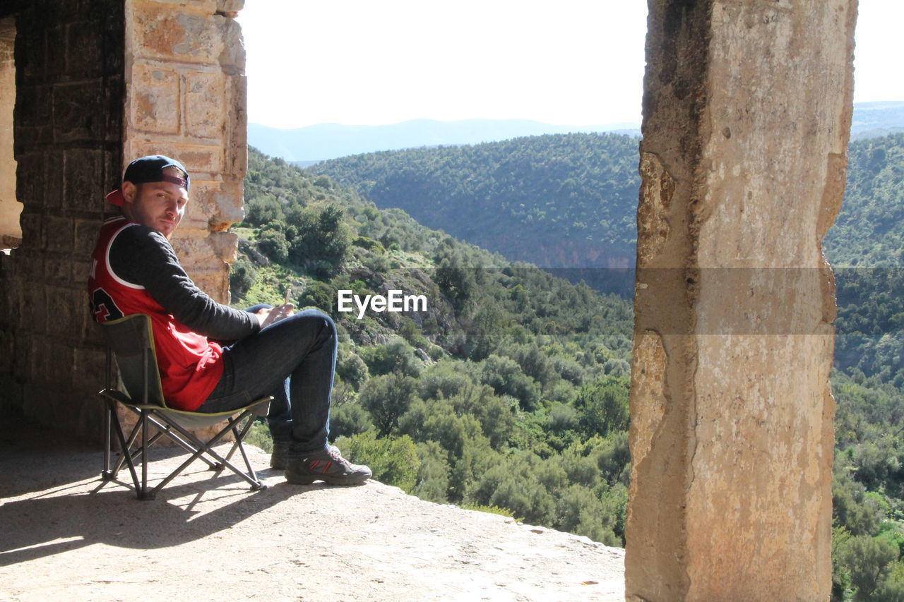 FULL LENGTH OF MAN SITTING ON SEAT AGAINST MOUNTAINS